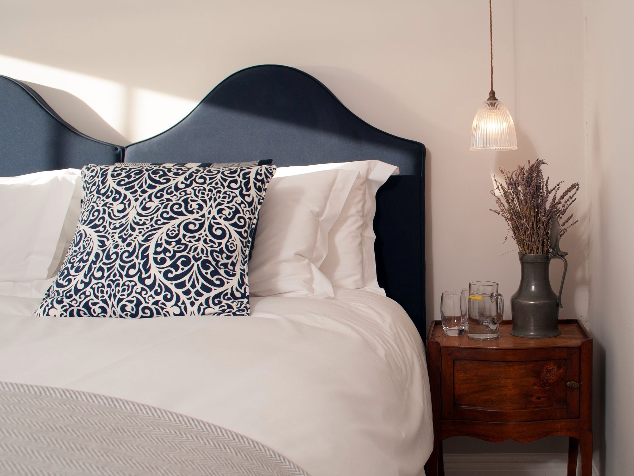 Una cama bien hecha con almohadas blancas y estampadas, un cabecero oscuro y una mesita de noche con un jarrón y vasos.