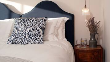 Una cama bien hecha con almohadas blancas y estampadas, un cabecero oscuro y una mesita de noche con un jarrón y vasos.