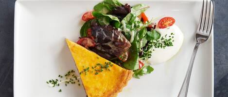 A white rectangular plate with a slice of golden-brown quiche, a mixed green salad with cherry tomatoes, and a dollop of white sauce, accompanied by a fork on a dark textured surface.