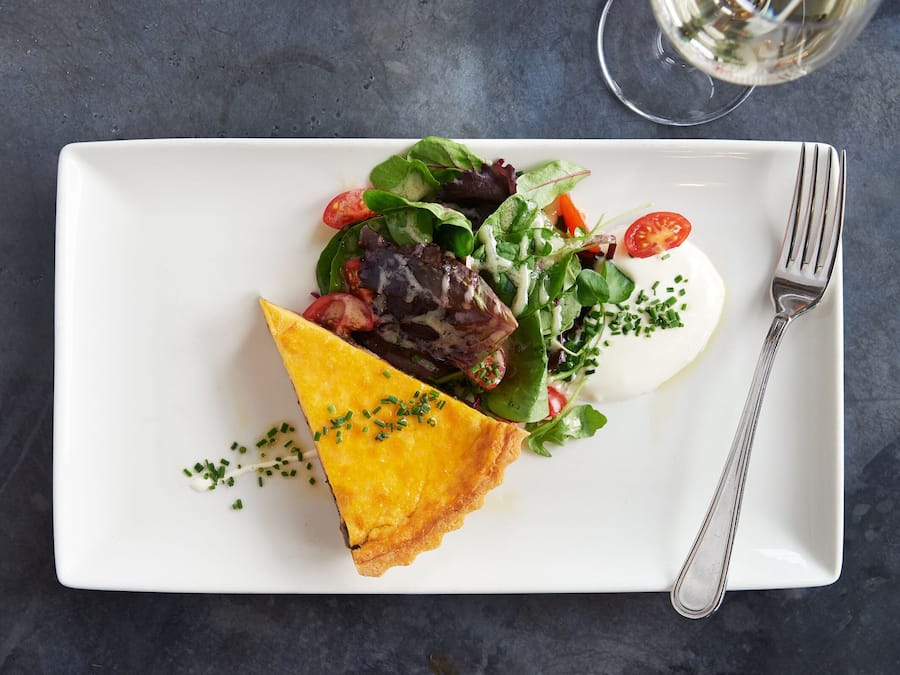 A white rectangular plate with a slice of golden-brown quiche, a mixed green salad with cherry tomatoes, and a dollop of white sauce, accompanied by a fork on a dark textured surface.