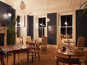 A dining room with a large table set for a meal, surrounded by chairs. There are multiple windows providing views of the city at night. - The House at Ramsgate (Ramsgate)