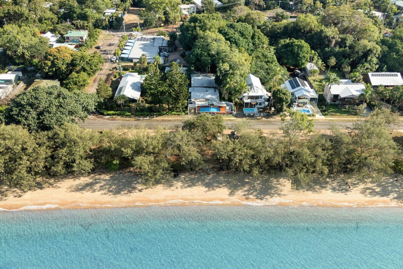 Vue aérienne d’un quartier résidentiel situé sur la côte, avec des maisons entourées d’une végétation luxuriante et une plage de sable.