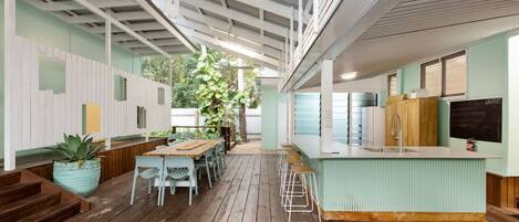 A spacious indoor area with a wooden floor, a long table with chairs, a bar area with stools, and a kitchen with a sink and cabinets.