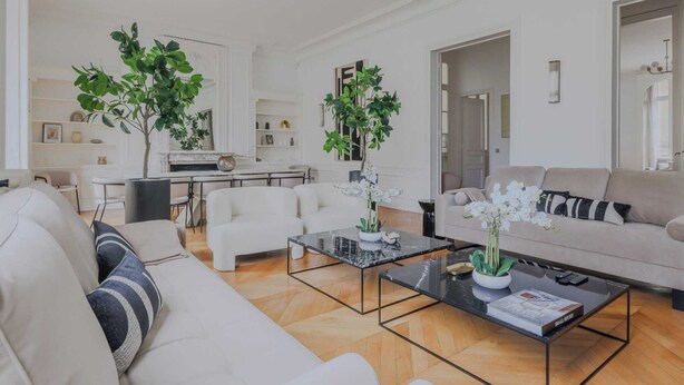Een moderne woonkamer met een bank, salontafel en twee potplanten. Achtergrond toont een eethoek met stoelen en een tafel.