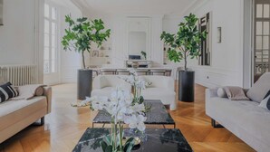 Een ruime woonkamer met een groot raam, twee potplanten, een salontafel en een bank.
