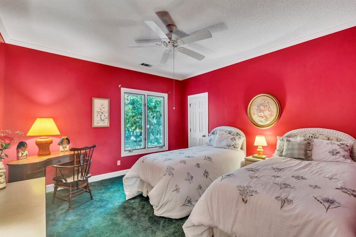 Une chambre avec deux lits, un bureau, une chaise, un ventilateur de plafond et une fenêtre donnant sur des arbres.