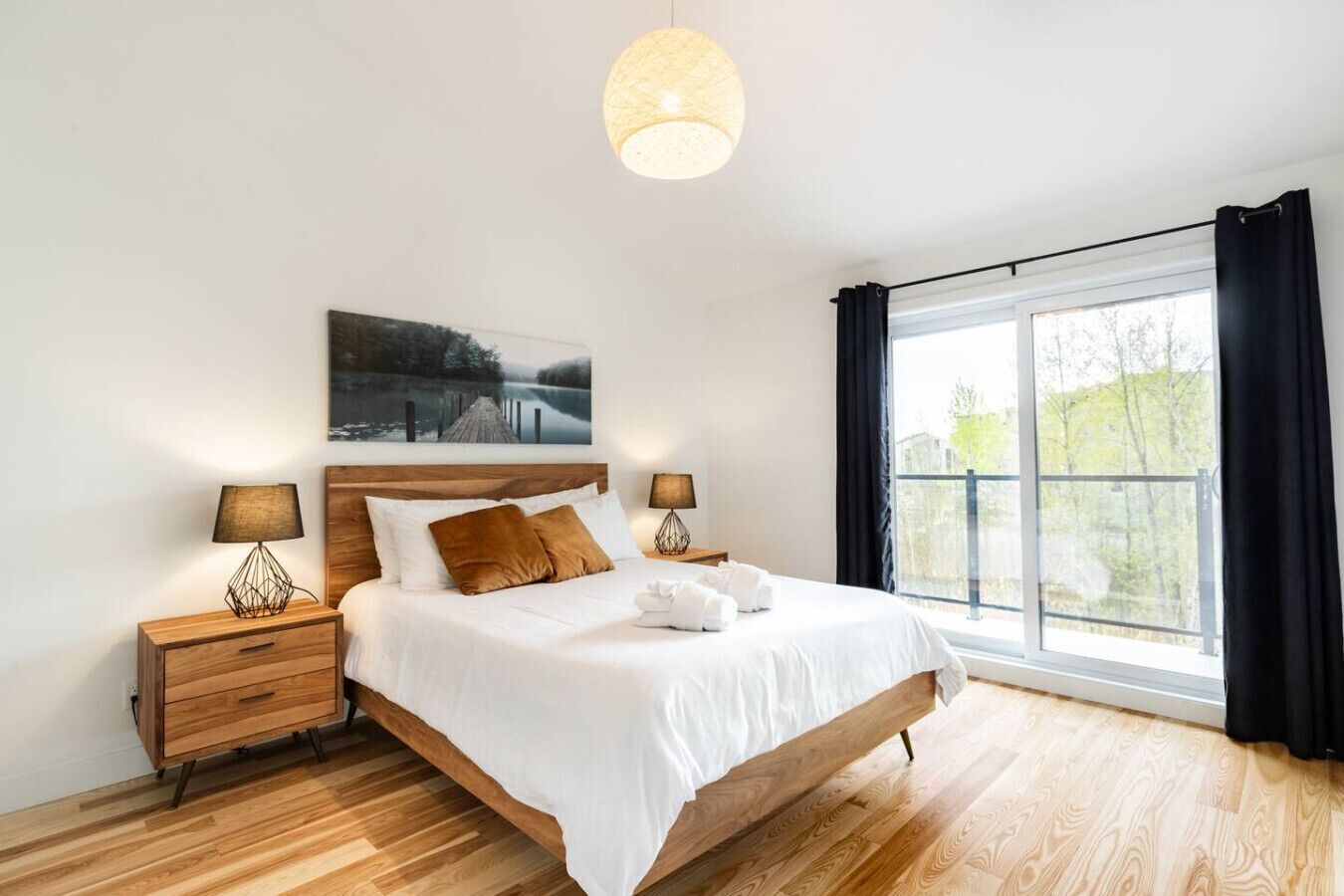 A modern bedroom with a wooden bed, two bedside tables with lamps, a large window with curtains, and a painting above the bed.