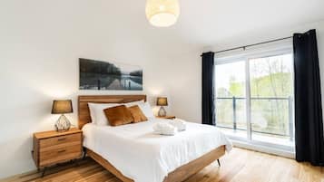 A modern bedroom with a wooden bed, two bedside tables with lamps, a large window with curtains, and a painting above the bed.