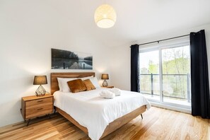 A modern bedroom with a wooden bed, two bedside tables with lamps, a large window with curtains, and a painting above the bed.