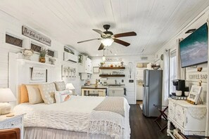 A cozy, well-lit bedroom with a bed, bedside table, and a ceiling fan. A kitchen area with a refrigerator and a coffee bar is visible in the background.