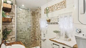A bathroom with a brick shower enclosure, wooden shelves with plants, a white sink, and a vintage-style vanity.