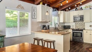 Una cocina con armarios blancos, una isla central, un microondas y una estufa. Hay dos taburetes de bar y una mesa de madera.