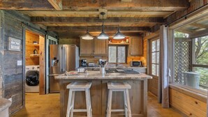 Cocina rústica de cabaña con paredes de madera, una isla central con taburetes, electrodomésticos de acero inoxidable y un ventanal amplio.