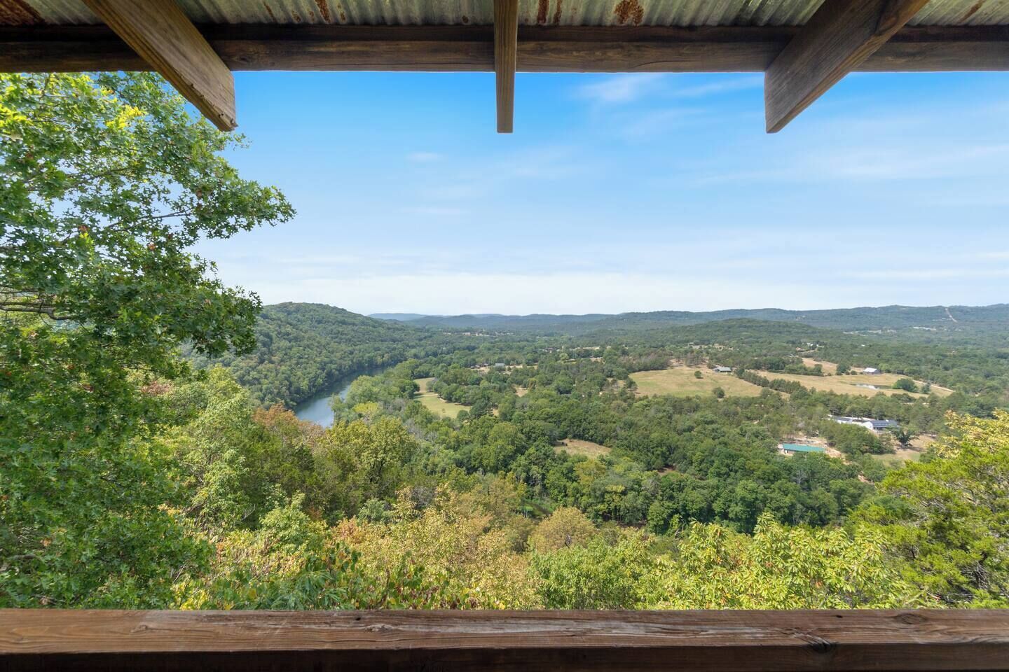 A view from a wooden balcony overlooking a river valley with dense forest and scattered buildings.