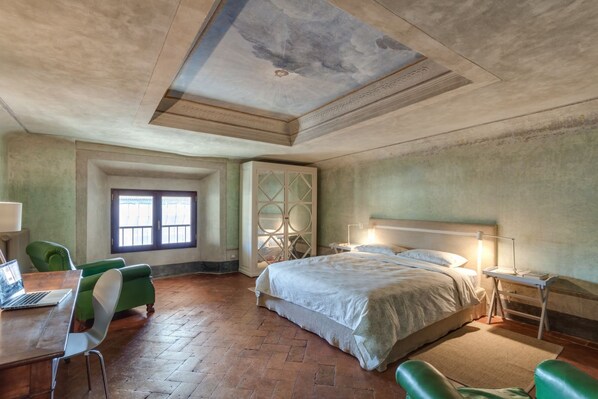 A bedroom with a large bed, a desk with a laptop, green chairs, a window, and a decorative ceiling.