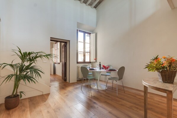 A spacious room with wooden flooring, a dining table with chairs, a potted plant, and a window providing natural light.