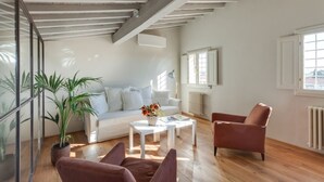Amplia sala de estar con un sofá blanco, una mesa de centro y un sillón marrón. Una ventana grande ofrece luz natural y vistas al exterior.