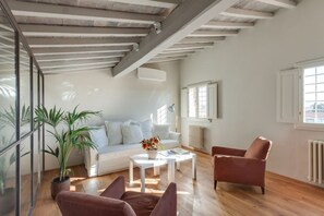 A spacious living room with a white sofa, a coffee table, and a brown armchair. There is a large window providing natural light and a view of the outside.