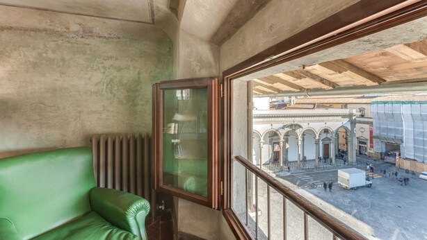 Una habitación con un banco de cuero verde, una ventana con marco de madera y vista a un patio exterior con arcos y un edificio en construcción.
