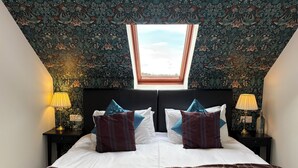 Un dormitorio con tragaluz, una cama con muchos cojines, dos mesitas de noche con lámparas y un papel tapiz estampado.