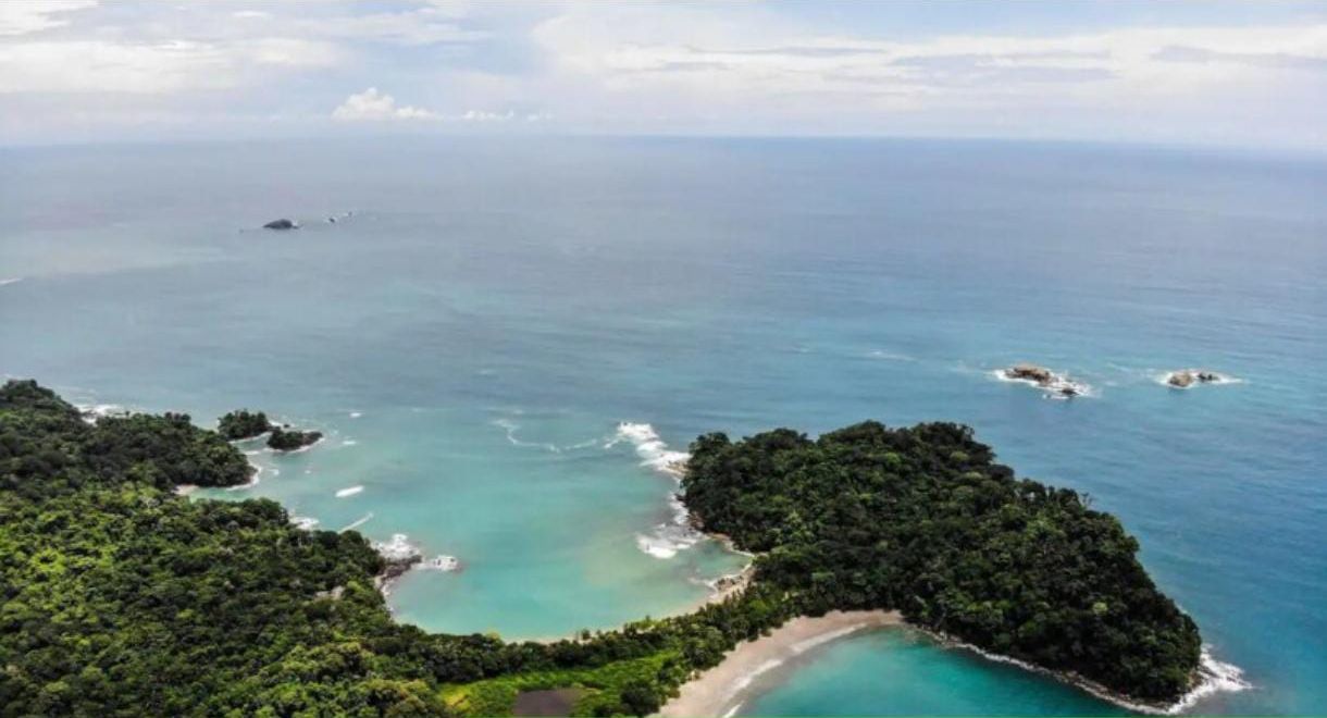 Vue a érienne d’une zone côtière avec des eaux turquoise limpides, une plage principale et une végétation luxuriante.