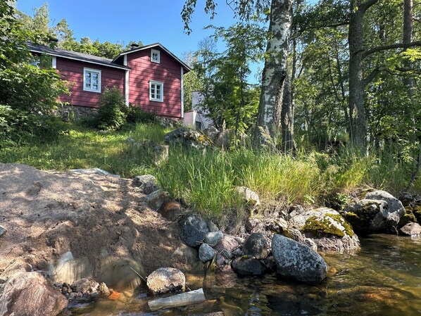 A red house with a clear stream in front, surrounded by rocks and greenery.
