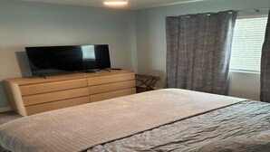 Une chambre à coucher comprenant un lit, une commode, un téléviseur et une fenêtre avec des rideaux.