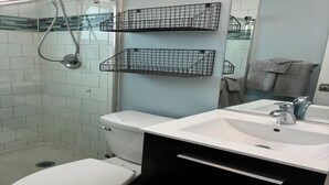 Une salle de bain avec des toilettes blanches, un lavabo avec un miroir et une douche. Deux paniers noirs en fil métallique sont accrochés au mur.