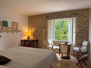 Een kamer met een bed, een tafel met stoelen, een lamp en een raam met uitzicht op groen.