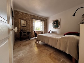 Ein Zimmer mit Bett, Sessel, Tisch, Fenster und einer Steinwand.