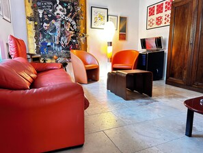 Een moderne woonkamer met een rode bank, oranje fauteuils, een kleine tafel en een wand versierd met diverse objecten en kunstwerken.