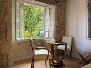 Une pièce avec un mur en pierre, une fenêtre donnant sur un paysage verdoyant, une chaise en bois, une petite table ronde et un présentoir à magazines.