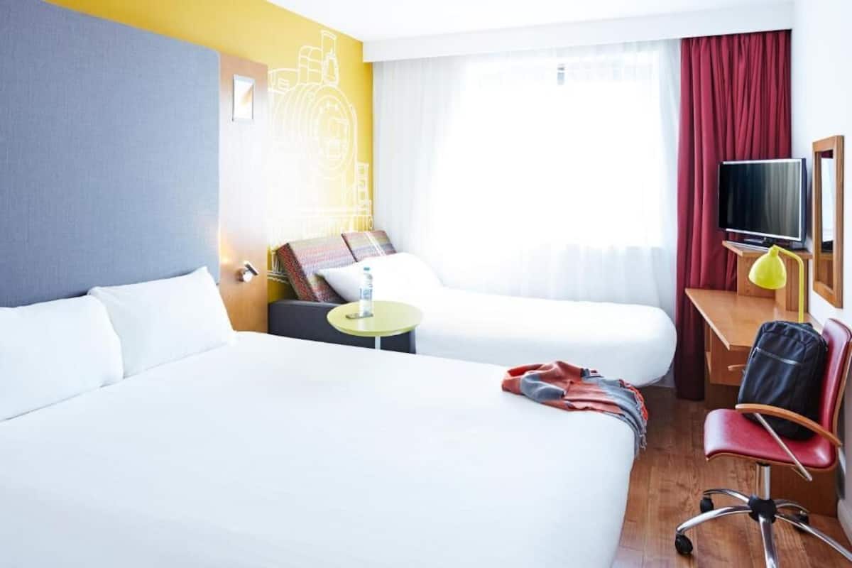 A hotel room with two beds, a desk with a TV, a chair, and a small table with a lamp.