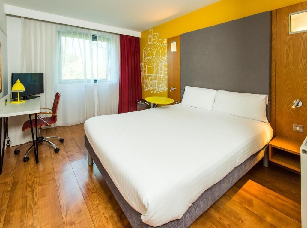 A hotel room with a large bed, a desk with a chair, a small round table, and a television.