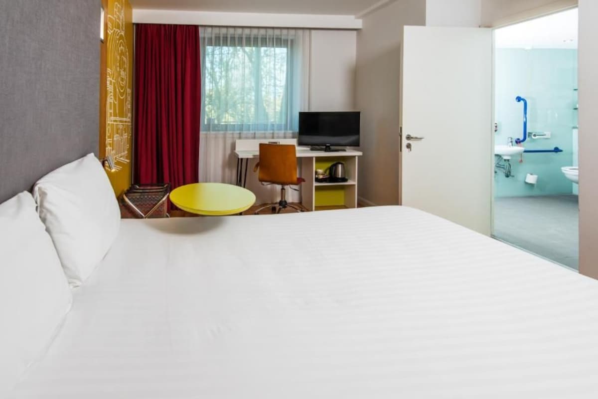 A hotel room with a bed, a desk with a TV, a chair, a small round table, and a bathroom visible through an open door.