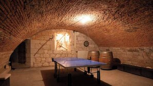 Ein Tischtennis-Tisch in einem Raum mit Backstein-Decke und Steinwänden, außerdem eine Dartscheibe und Holzfässer.