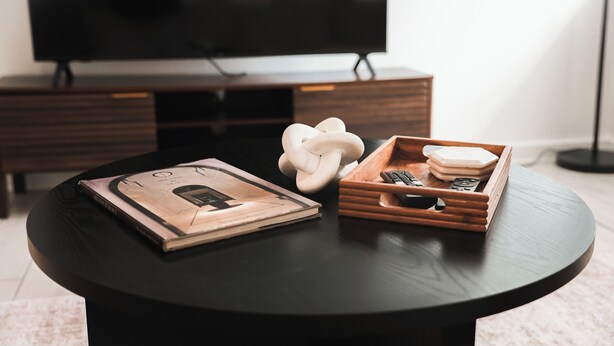 Een ronde zwarte tafel met een tijdschrift, een witte sculptuur en een houten dienblad met een afstandsbediening en boeken.