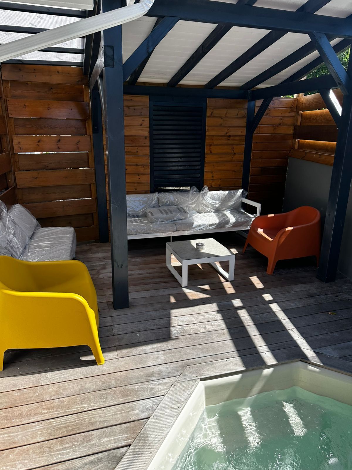 A covered outdoor seating area with wooden benches, a yellow chair, a white table, and a hot tub.