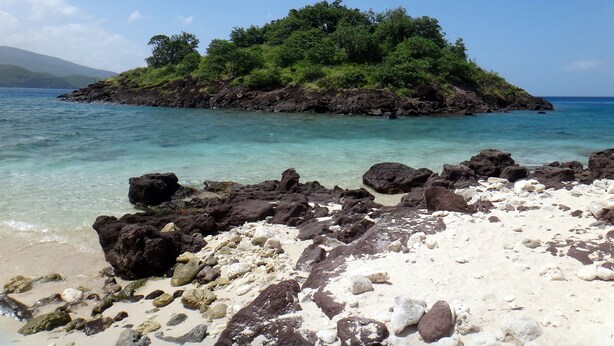 Una playa rocosa con agua turquesa cristalina y una pequeña isla con vegetación verde exuberante al fondo.