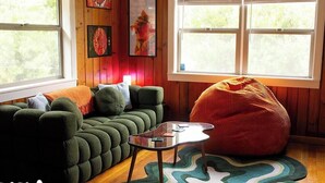 Ein gemütliches Zimmer mit einem grünen Sofa, einem roten Sitzsack, einem kleinen Tisch und einem Fenster mit Blick auf Grünflächen.