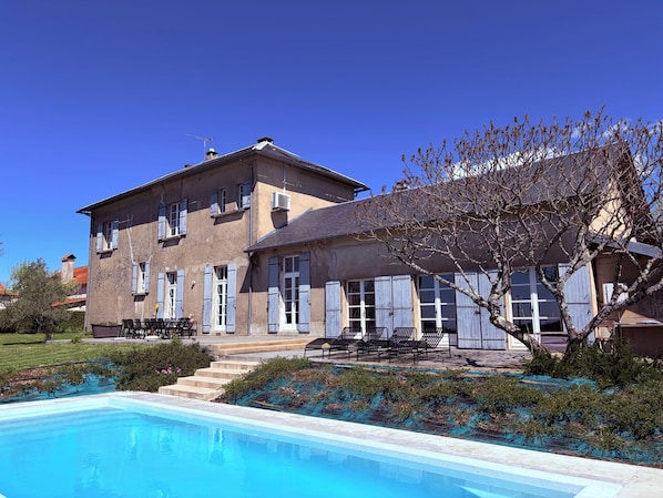 A two-story building with a swimming pool, outdoor seating, and a tree in the foreground.