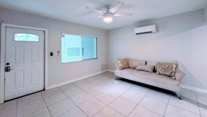 Ein geräumiger Raum mit weißer Tür, einem Fenster mit Jalousien, einem Deckenventilator und einem beige gefärbten Sofa.