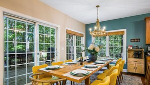 A dining area with a long table set for six, surrounded by yellow chairs, a chandelier, and large windows with blinds.