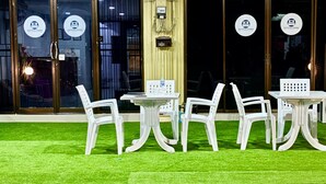 Un espace de détente intérieur avec des chaises et des tables en plastique blanc, un sol recouvert d’un gazon artificiel vert et une fenêtre donnant sur l’extérieur.