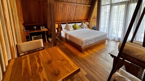 A wooden-themed bedroom with a bed, bedside table, chair, and a desk with a lamp.