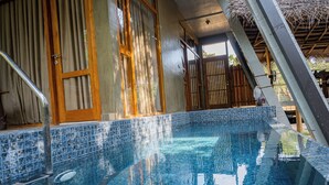 Una piscina azul cristalina con bordes de azulejos, puertas de madera y una escalera metálica que da acceso al interior.