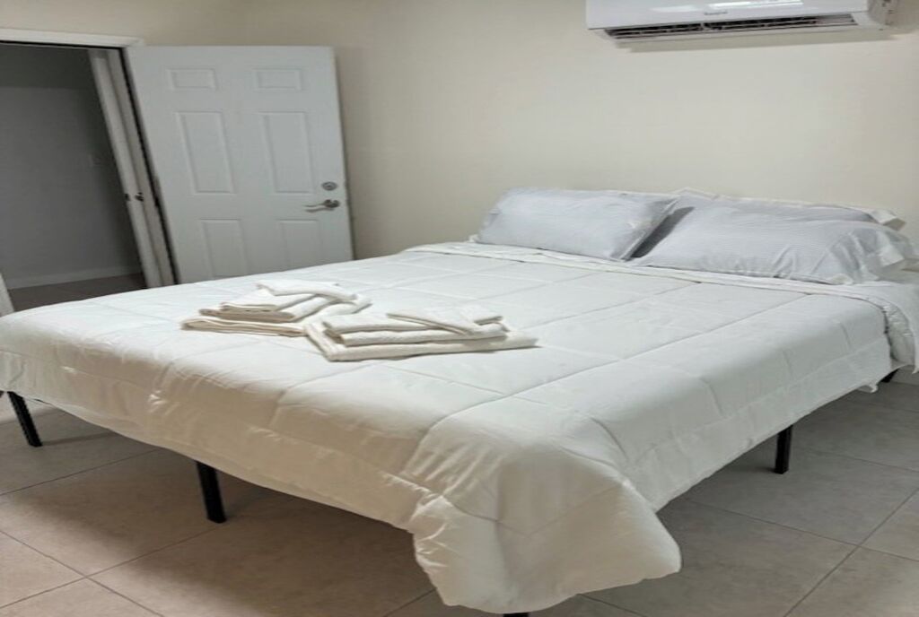 A neatly made bed with white linens and towels, a white door, and a white air conditioning unit on the wall.