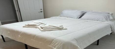 A neatly made bed with white linens and towels, a white door, and a white air conditioning unit on the wall.