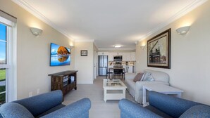 Ein Wohnzimmer mit blau gemusterten Sofas, einem weißen Couchtisch, einem Fernseher und einer Küche im Hintergrund.