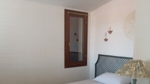 A small, single-bed room with a wooden headboard, a bedside table with a lamp, a window with a wooden frame, and a wall-mounted light fixture.